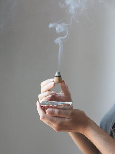 woman's hands holding an abalone shell and a palo santo aromatic incense cone burning with smoke billowing