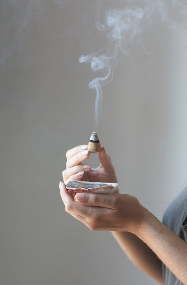 A person holds a smoking palo santo aromatic incense cone above a hand holding an abalone shell incense burner