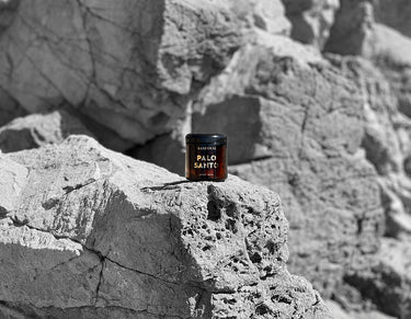 An amber glass jar with gold words that say sandoval palo santo aromatic incense, as pictured on a rock cliff side in Malibu California