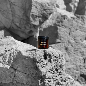 An amber glass jar with gold words that say sandoval palo santo aromatic incense, as pictured on a rock cliff side in Malibu California